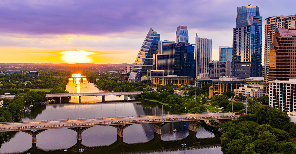 A stunning drone aerial view captures the Austin, Texas downtown skyline bathed in golden and purple sunset light, with the Colorado River (Lady Bird Lake) stretching beneath the Congress Avenue Bridge. Kayakers and paddleboarders dot the calm water below while crowds gather along the bridge to watch the spectacular summer sunset. The distinctive pyramid-shaped Frost Tower and surrounding modern glass skyscrapers reflect the warm evening light against a dramatic purple sky.