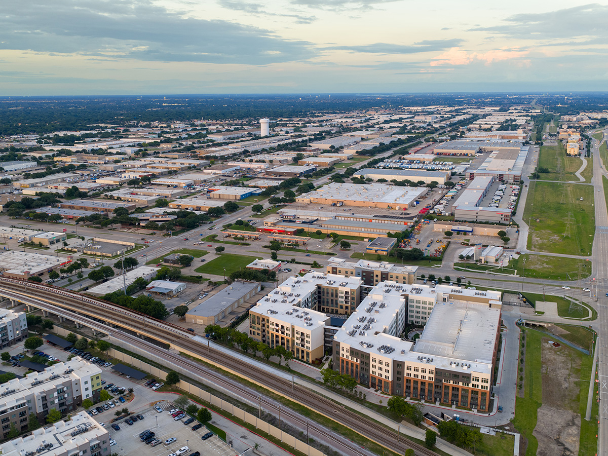 Drone aerial photograph showing a large-scale mixed-use development with modern apartment buildings, retail centers, and extensive highway infrastructure. The image captures urban sprawl and planned community development in a metropolitan area during golden hour lighting conditions.