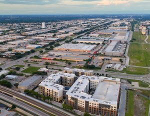 Drone aerial photograph showing a large-scale mixed-use development with modern apartment buildings, retail centers, and extensive highway infrastructure. The image captures urban sprawl and planned community development in a metropolitan area during golden hour lighting conditions.