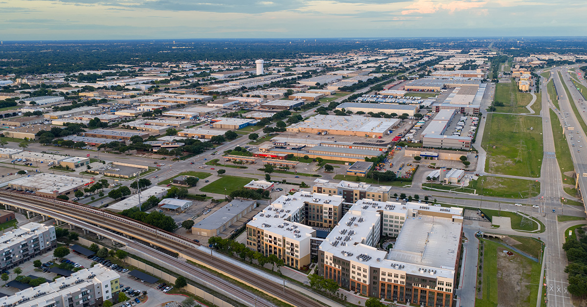 Drone aerial photograph showing a large-scale mixed-use development with modern apartment buildings, retail centers, and extensive highway infrastructure. The image captures urban sprawl and planned community development in a metropolitan area during golden hour lighting conditions.