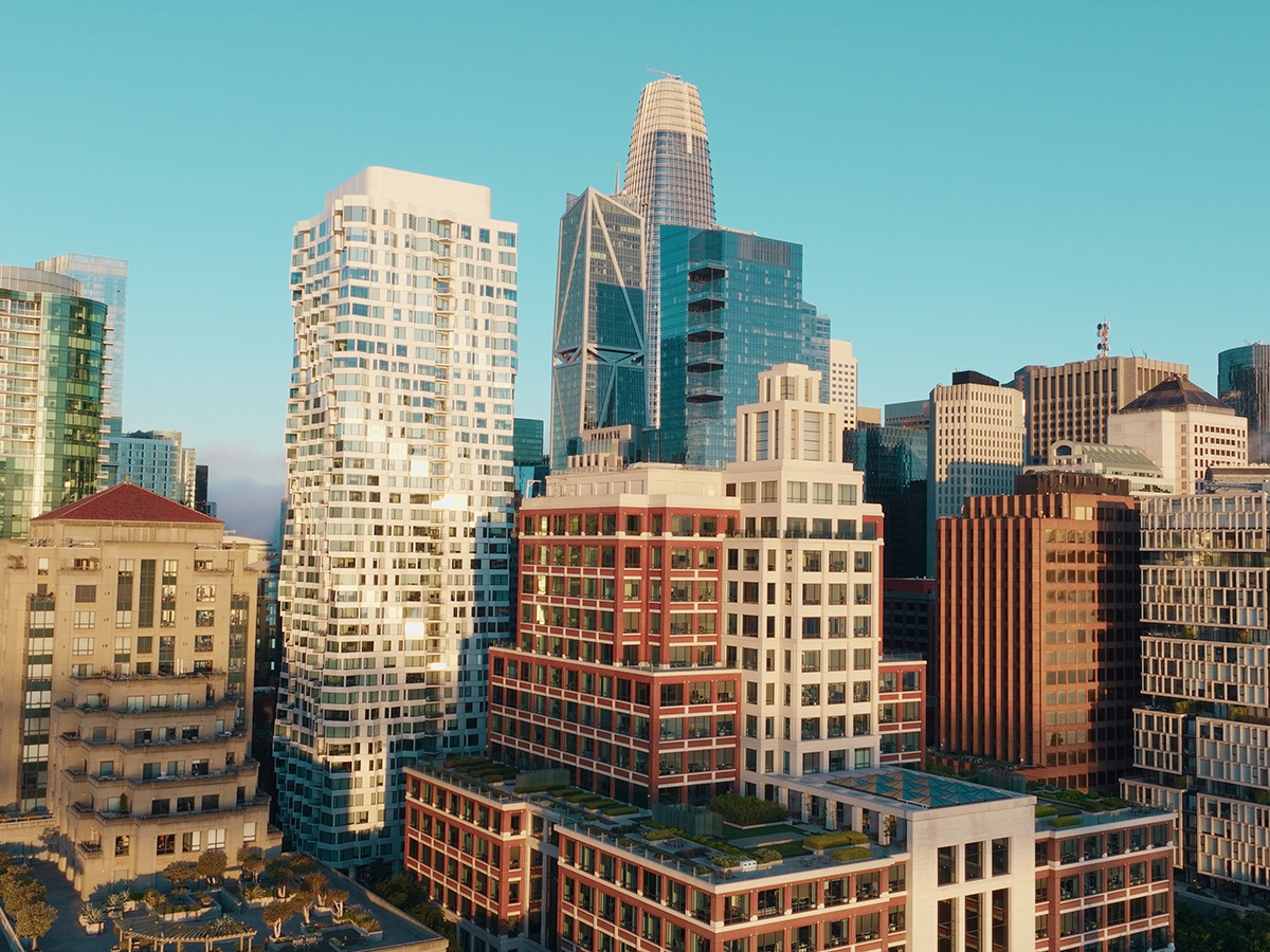 San Francisco is showcased in this stunning aerial view of its dense urban skyline. Modern skyscrapers, diverse architecture, and rooftop gardens are bathed in warm golden hour sunlight against a clear blue sky.