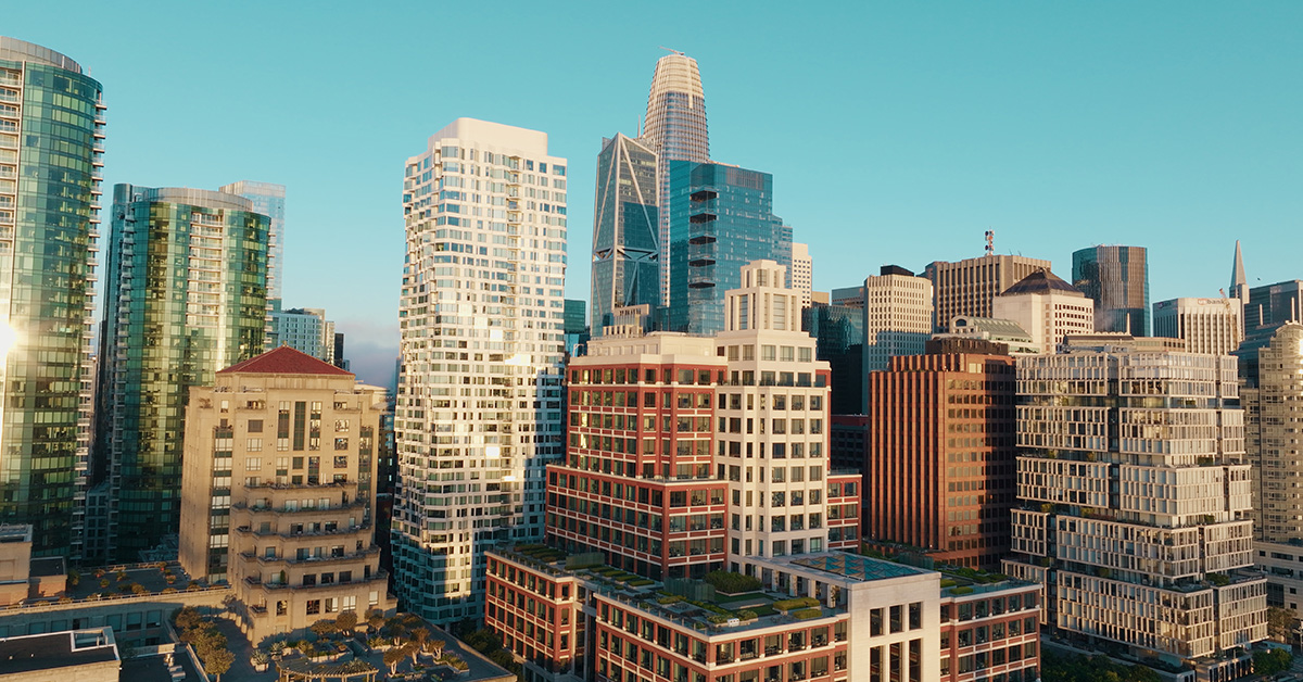 San Francisco is showcased in this stunning aerial view of its dense urban skyline. Modern skyscrapers, diverse architecture, and rooftop gardens are bathed in warm golden hour sunlight against a clear blue sky.