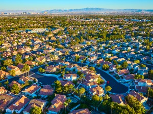 Aerial view of a serene suburban neighborhood in Las Vegas, Nevada. Warm golden hour light highlights red-tiled rooftops and green trees, with a city skyline and mountains in the distance.