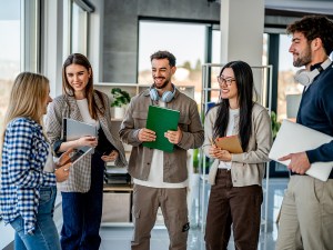Group of happy young adult students smiling and talking, holding laptops and books inside a modern university building