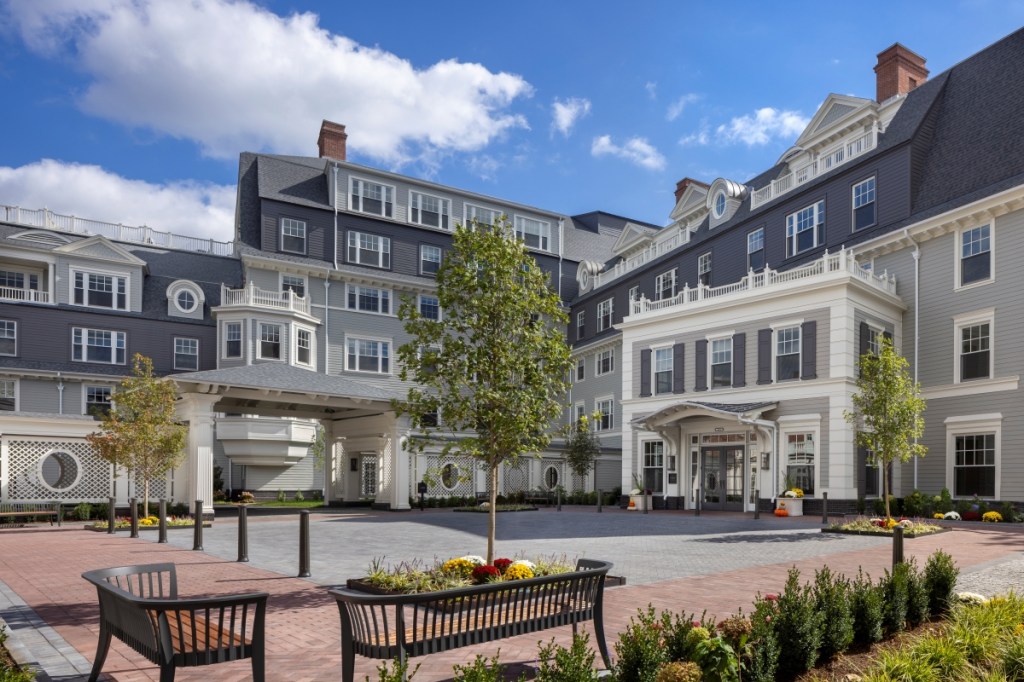 Exterior view of The Newbury of Brookline, showcasing its classic design and welcoming entrance surrounded by greenery.