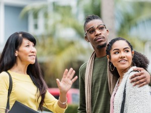 A mature mixed race Caucasian and Chinese woman standing outdoors, carrying a leather file organizer, residential buildings out of focus in the background. She is a real estate agent showing a home to a young African-American couple.
