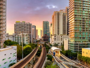 Public train system travels along elevated railroad in Miami downtown office district at sunset, with tall commercial and residential skyscrapers surrounding the tracks.
