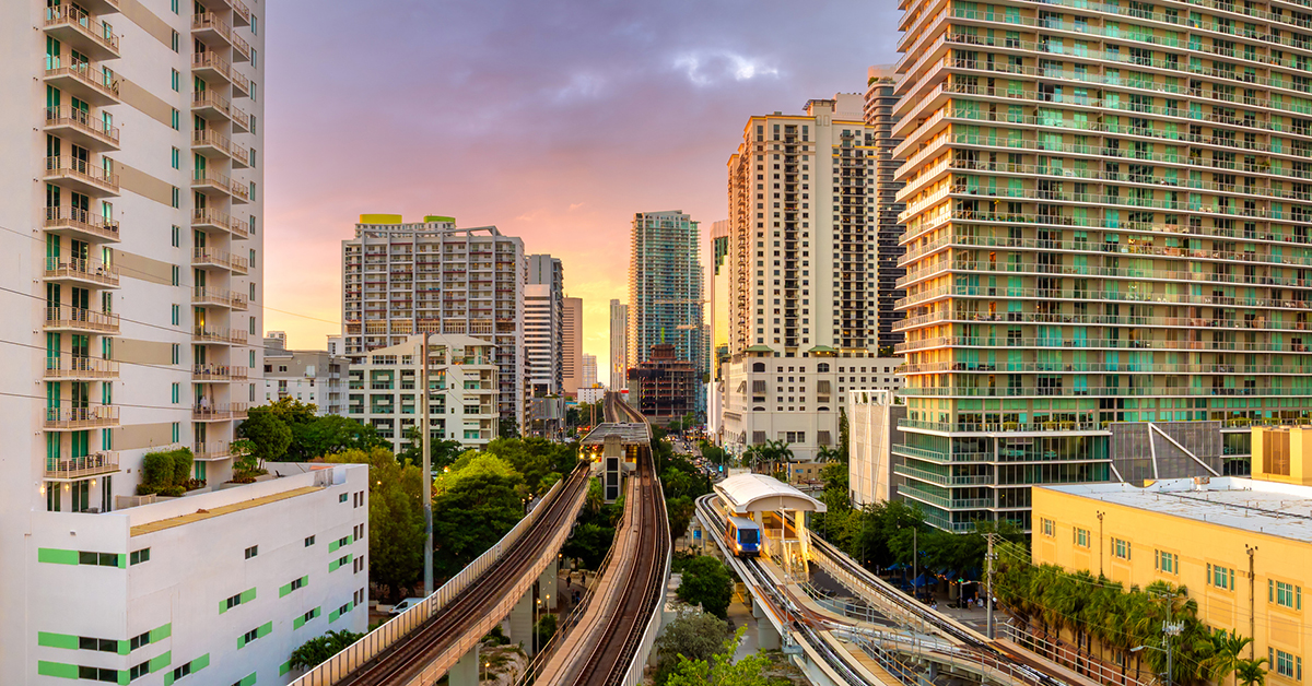 Public train system travels along elevated railroad in Miami downtown office district at sunset, with tall commercial and residential skyscrapers surrounding the tracks.